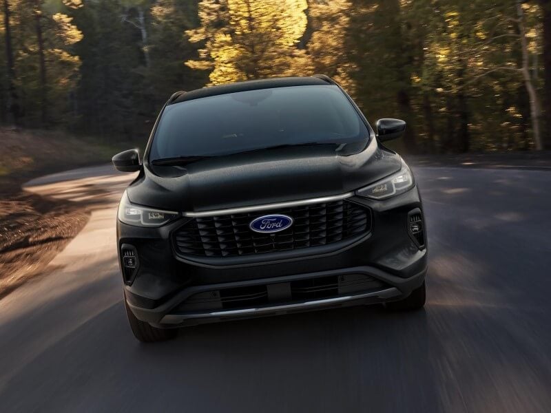 A black 2026 Ford Escape SUV driving on a winding road through a sunlit forest.